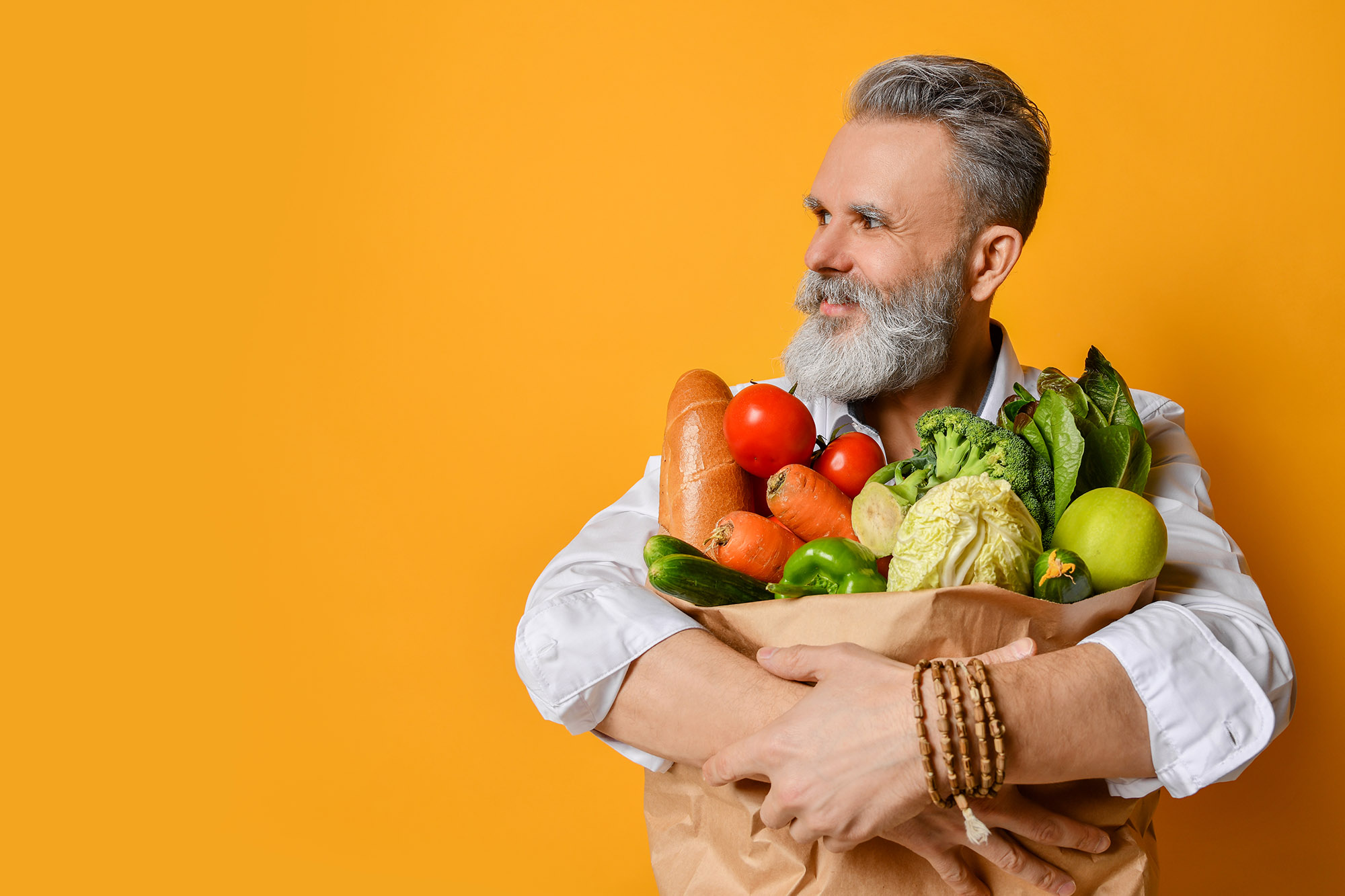 Phone conversation. Positive senior aged man holding groceries on a yellow background